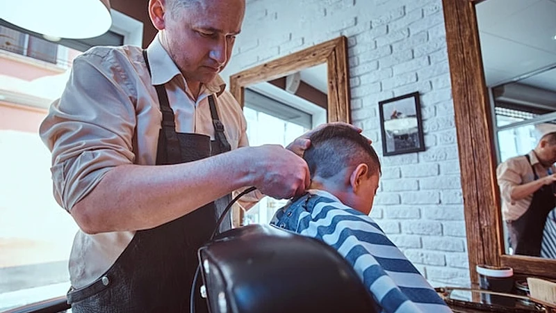 Small school boy is getting trendy haircut from expirienced barber at modern barbershop.