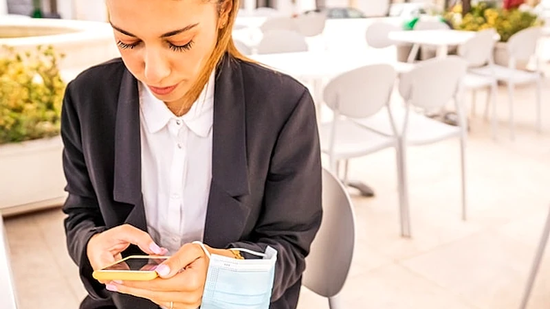 Young woman self entrepreneur texting on smartphone sitting at bar dehors with holding protective face mask in hand. Freelance girl taking breakfast at hotel cafe using technology. New normal habits
