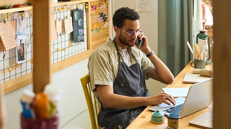 Young male clerk in apron calling clients and looking through online orders