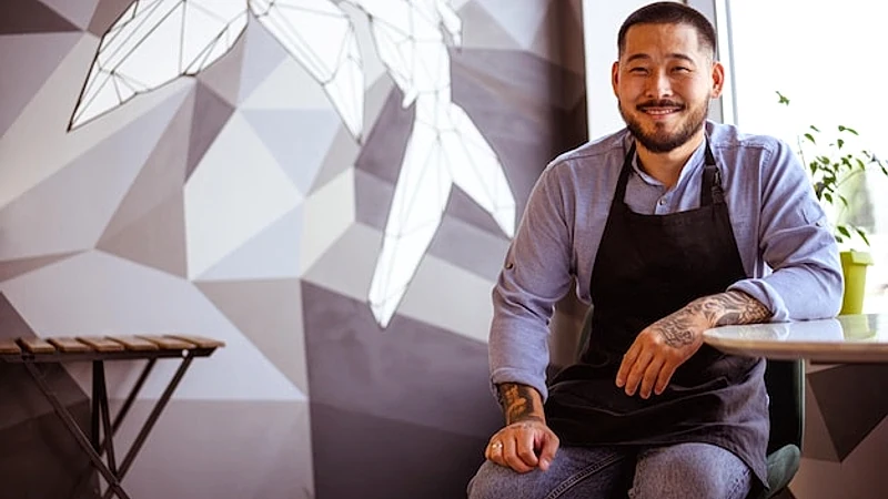 Joyful asian owner in apron and shirt smiling at camera in cafe