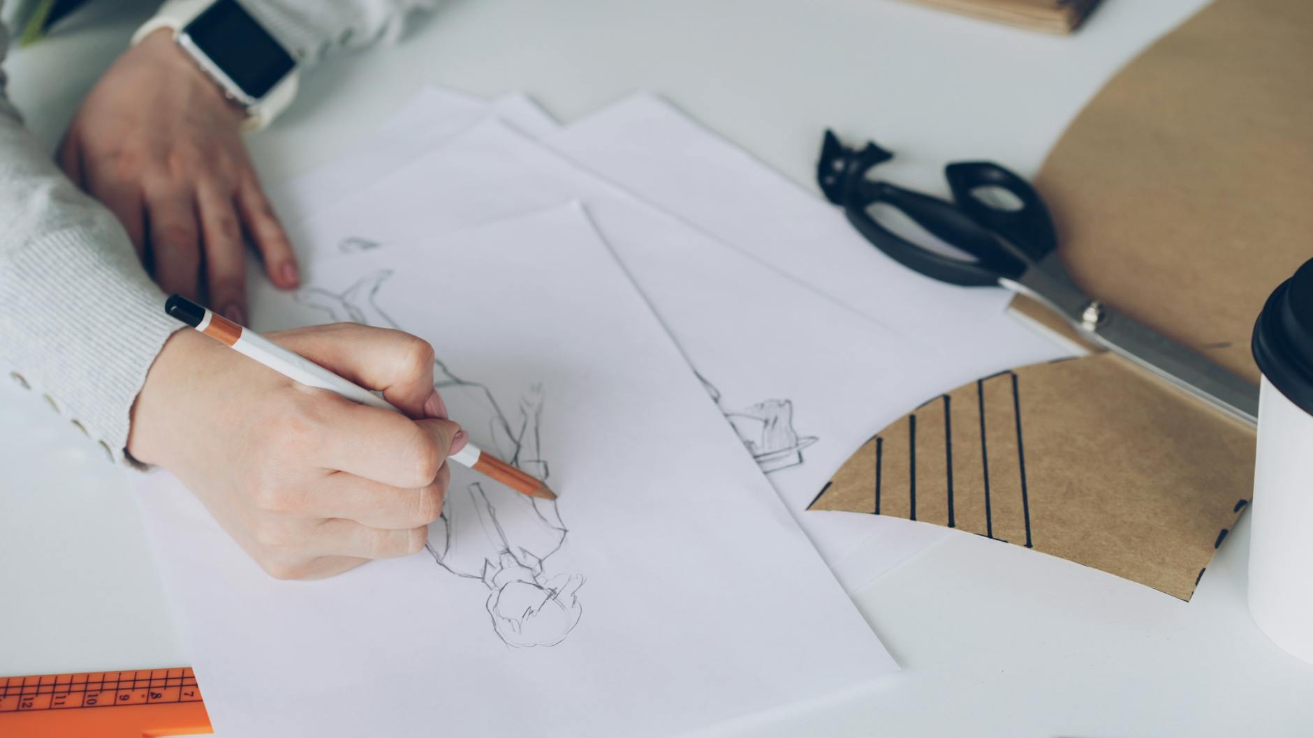 Artist's hand sketching fashion designs with paper and tools in view.