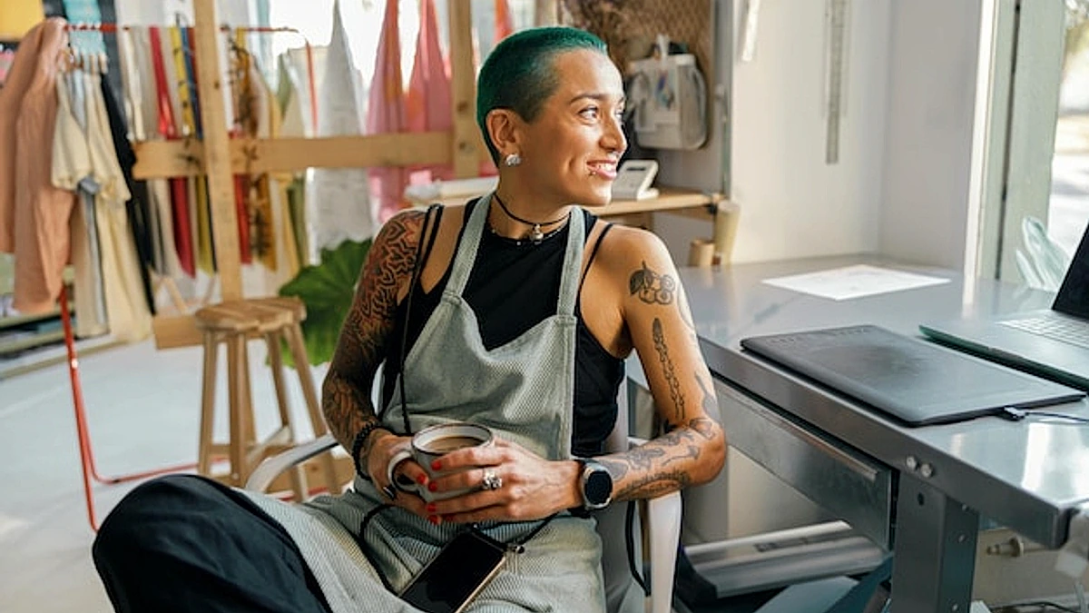 Female worker sitting with coffee near desk with laptop in design studio and looks camera