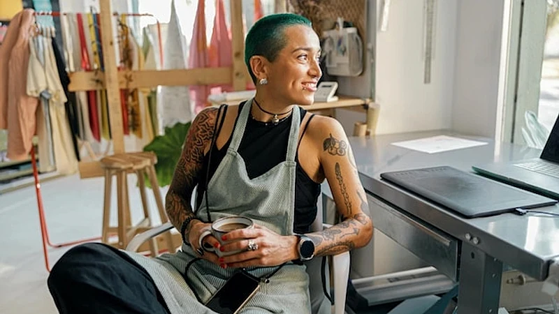 Female worker sitting with coffee near desk with laptop in design studio and looks camera