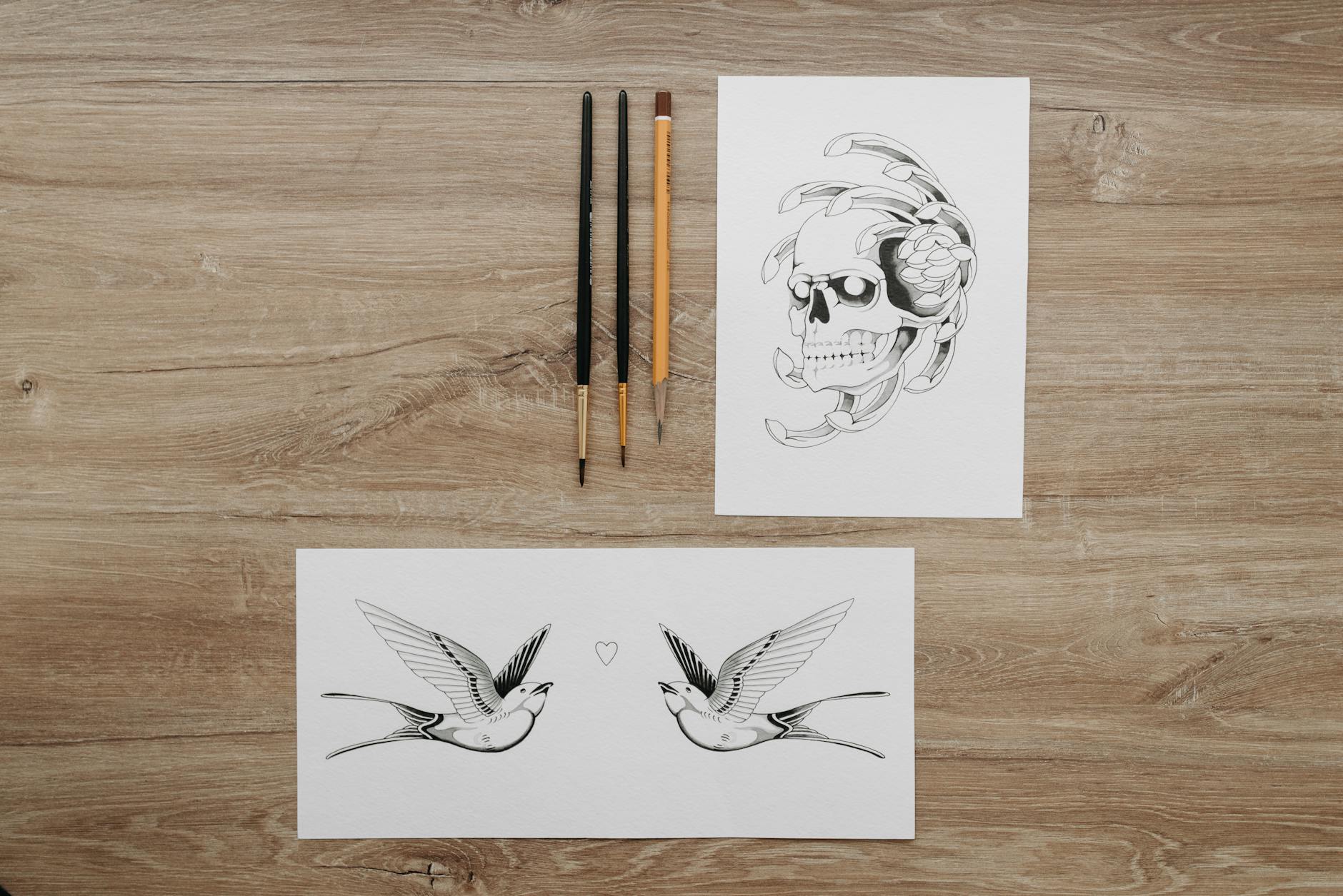 Flat lay of tattoo design sketches featuring a skull and swallows with art brushes on a wooden table.