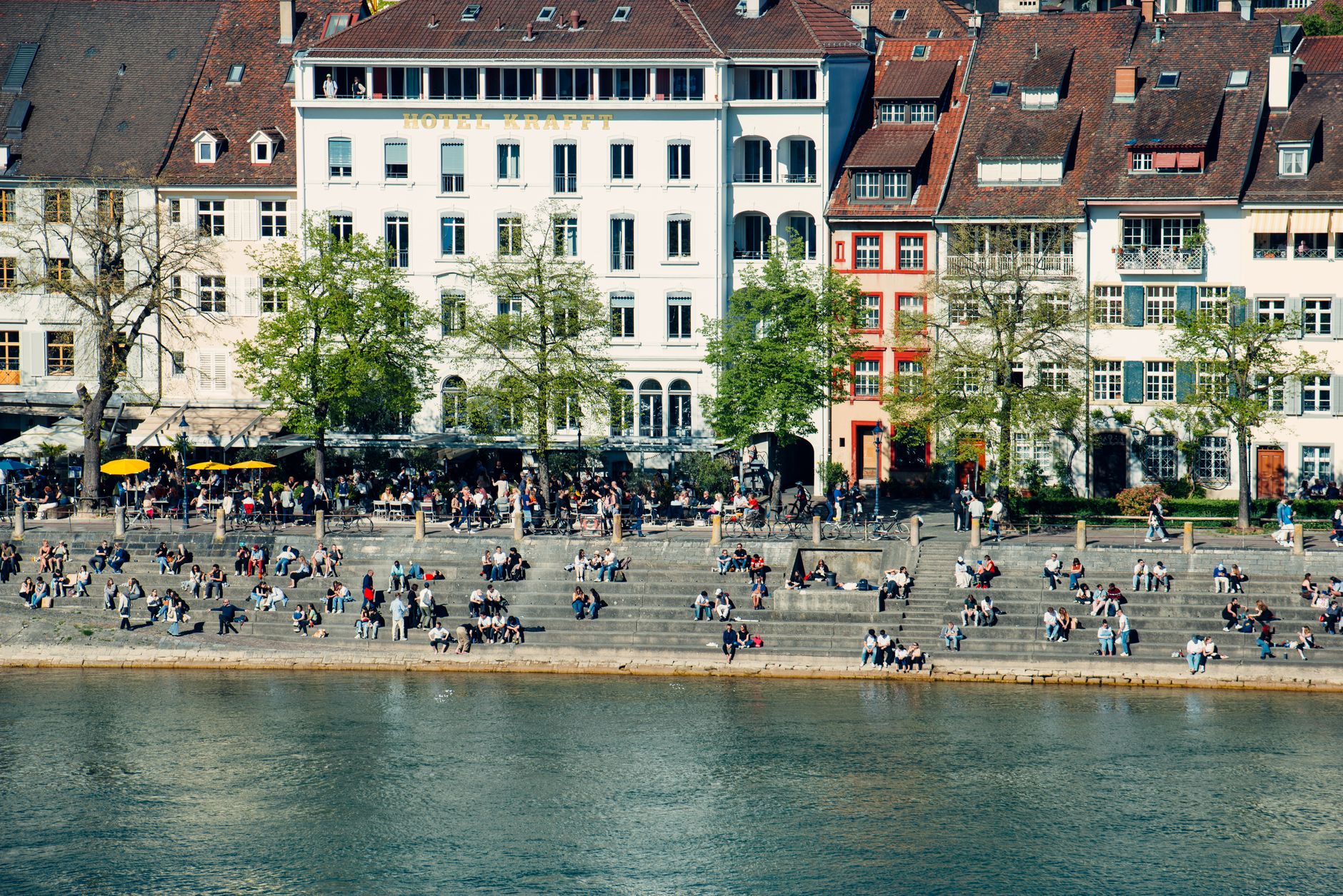 People relax and socialize by the Rhine River in Basel, showcasing vibrant city life.