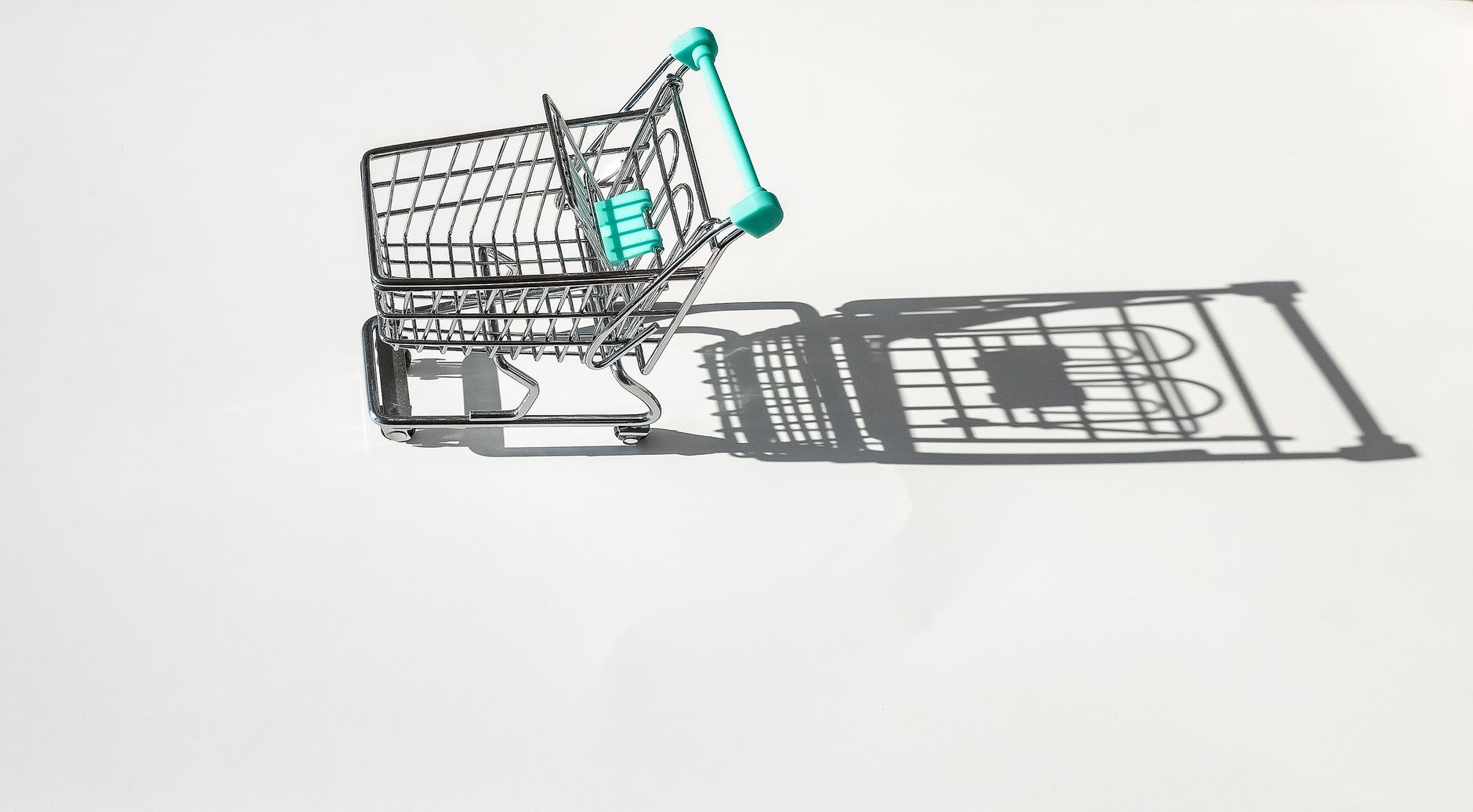 A minimalist image of a small shopping cart casting a shadow on a white surface, captured from above.