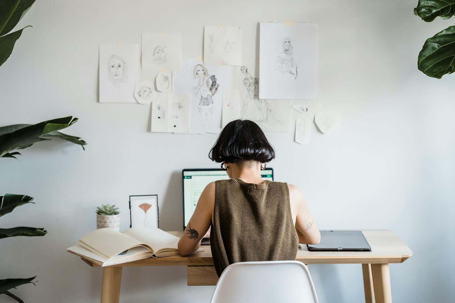 Back view of unrecognizable female artist with short dark hair in casual clothes sitting at table and working on laptop in light room with paints on wall and green plants
