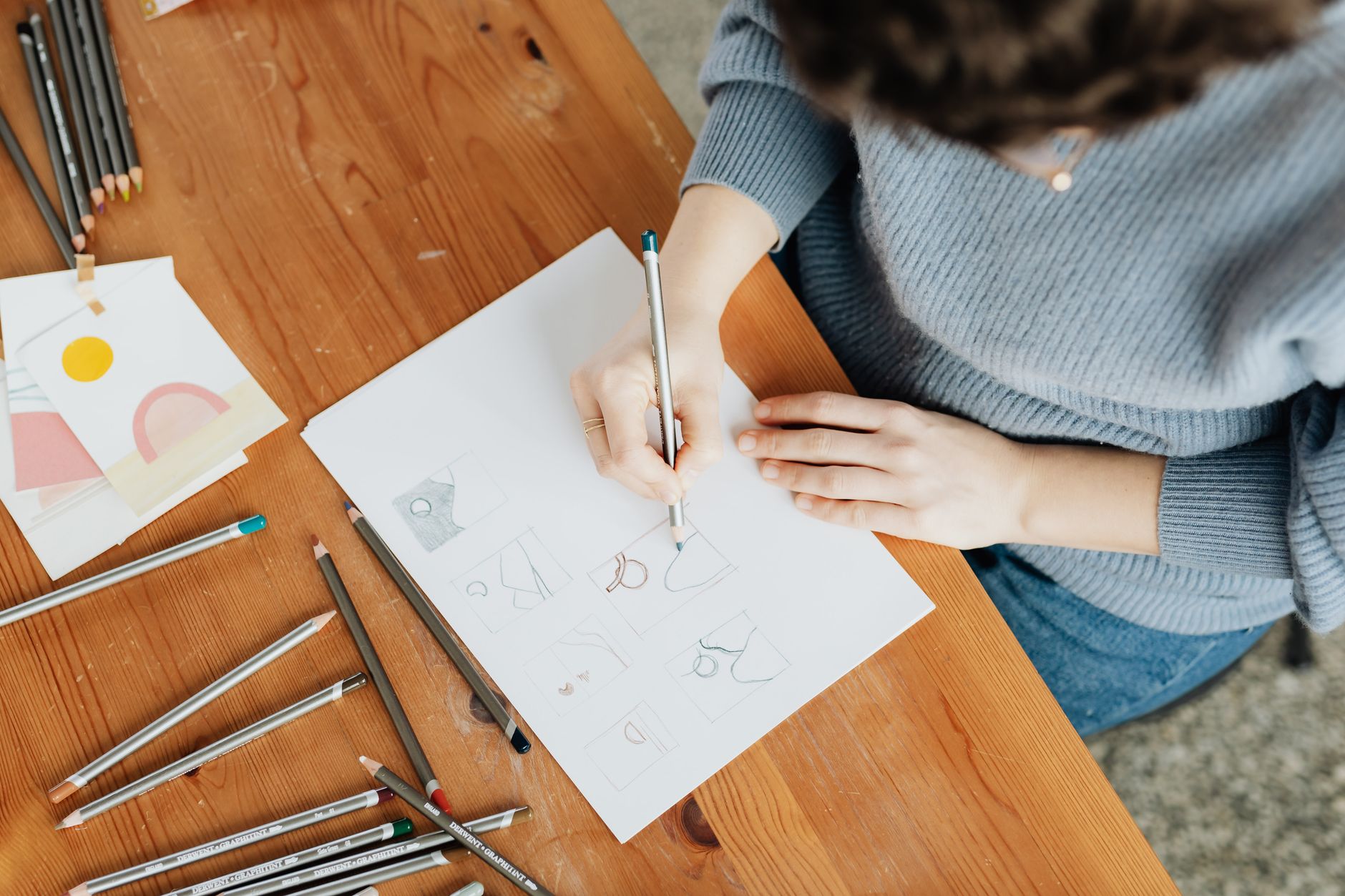 Top view of crop anonymous female illustrator in sweater and jeans with eyeglasses making sketch with pencil on paper while sitting at wooden desk at work
