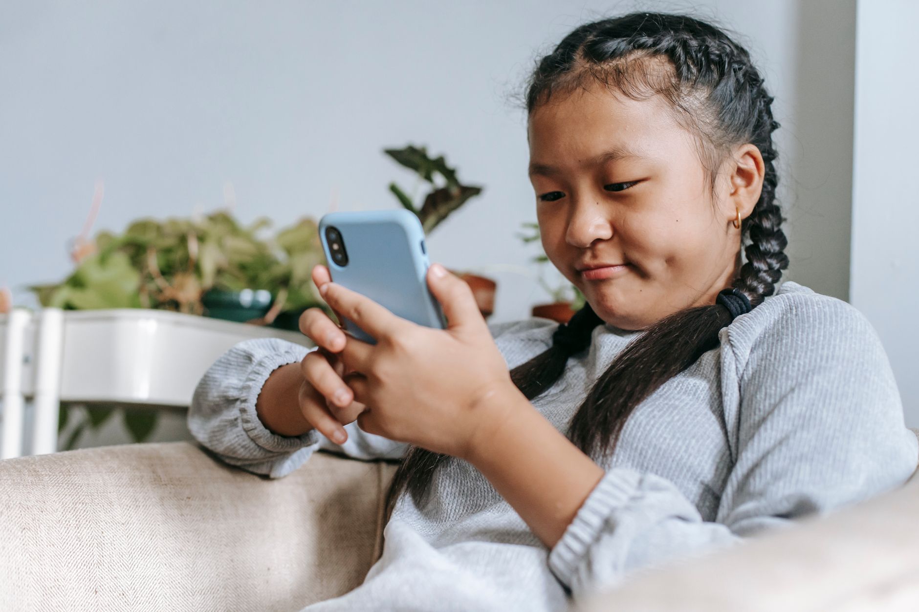 Focused ethnic girl in casual outfit sitting on comfortable armchair and browsing internet on smartphone