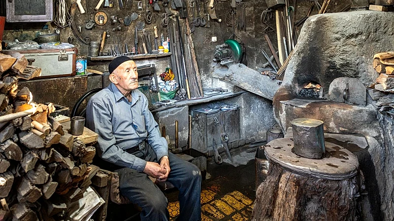 An elderly blacksmith sitting in a cluttered workshop surrounded by tools and firewood.